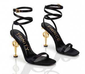 Black high-heeled sandals with gold accents and 'Trinity' and 'Prince' branding on a white background.